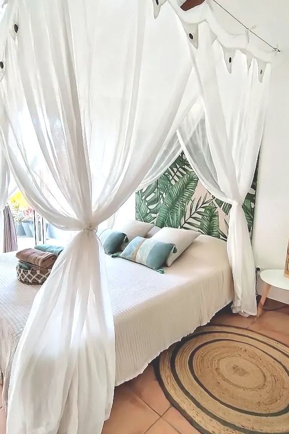 A cozy bedroom is equipped with a spacious bed draped in flowing white curtains, enhancing the tranquil ambiance. Decorative green accents adorn the wall behind the bed, which is complemented by two soft pillows. A round woven rug adds texture to the tiled floor.