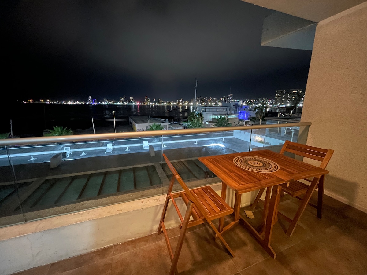 An outdoor terrace features a wooden table and two folding chairs. The railing provides an unobstructed view of the illuminated coastline and city skyline at night. Soft ambient lighting enhances the tranquil setting, inviting relaxation while enjoying the sea breeze.