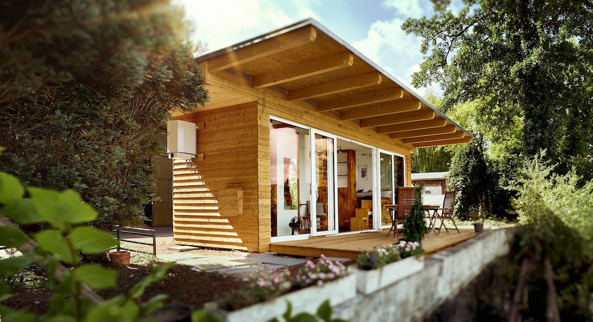 A modern wooden cabin features large glass doors that open to a welcoming porch area. Surrounded by greenery, the structure is complemented by a wooden overhang. Tables and chairs invite relaxation just outside, while bright natural light enhances the inviting atmosphere.