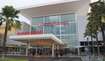 The exterior of Metromall is shown, featuring a modern design with large glass windows and an inviting entrance. The name 'Metromall' is prominently displayed in red. Palm trees are positioned around the entrance, adding greenery to the scene.