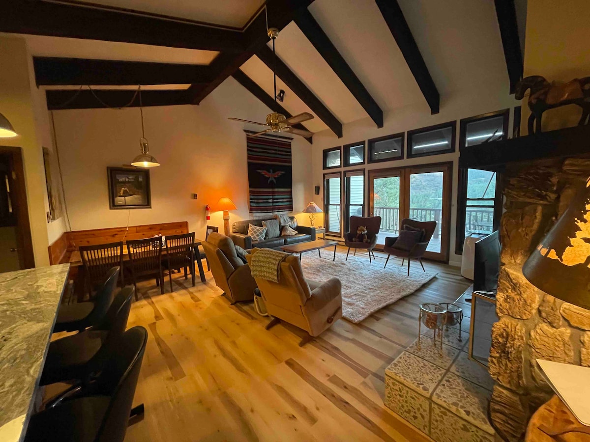 A spacious living area features high, exposed beam ceilings and large windows that provide natural light and mountain views. A cozy seating arrangement includes an area rug, comfortable chairs, and a sofa, with a dining table nearby and a fireplace adding warmth to the room.