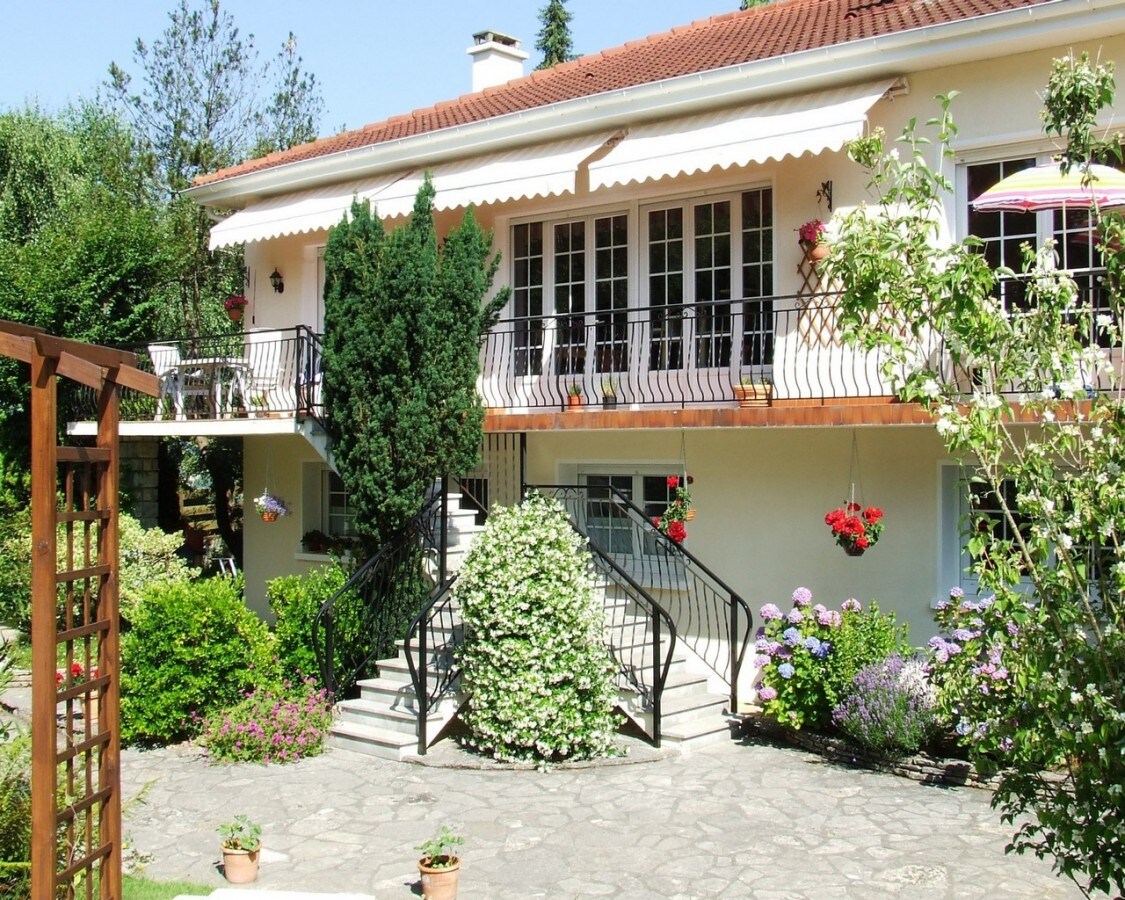 A charming two-story building features a welcoming entrance with a light-colored facade and a red roof. Flanking the entrance are flowering plants and greenery, enhancing the natural surroundings. A staircase leads up to a balcony adorned with colorful flowers and seating, providing a serene outdoor space.