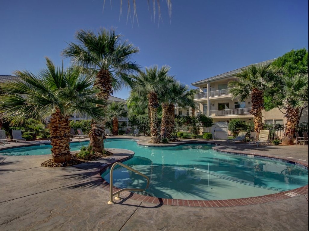 A tropical pool area is surrounded by palm trees and features a winding pool with a textured stone deck. Two lounge chairs are positioned nearby, offering an inviting space for relaxation. Views of the adjacent building and clear blue skies are visible in the background.