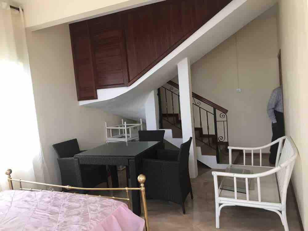 A comfortable living area is highlighted by a dark dining table surrounded by four chairs, complemented by a white seating area. A staircase with a wooden railing leads to the upper level, while light filters through the nearby window, enhancing the open space.