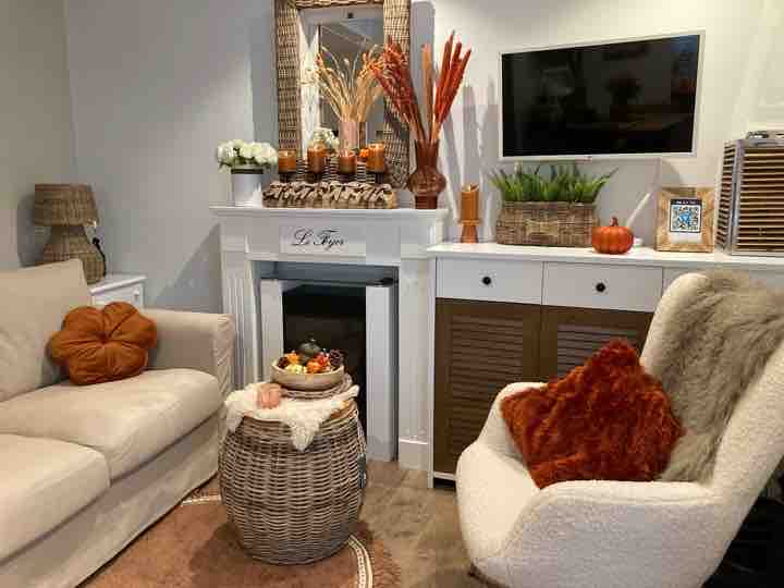 A cozy living area is featured with a light-colored sofa and a cream-colored armchair. A wicker coffee table holds decorative items, while a fireplace adds warmth. Framed mirrors and autumn-themed decorations enhance the welcoming ambiance, with greenery in woven containers subtly displayed.