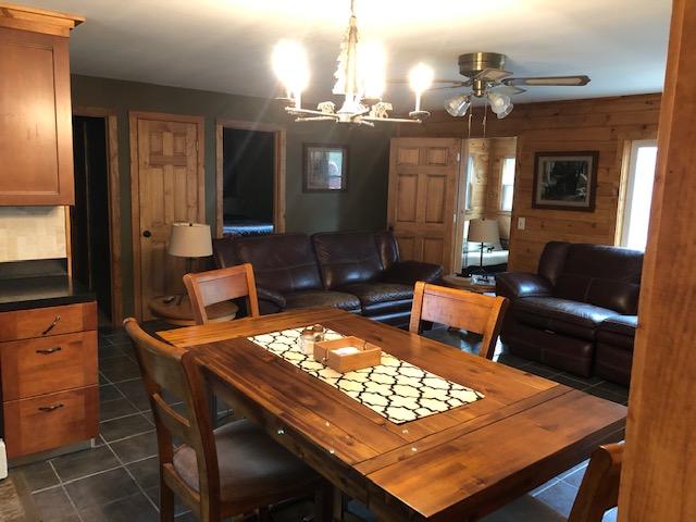 A cozy dining area features a wooden table surrounded by four chairs, with a decorative centerpiece. In the background, a comfortable living space is visible, showcasing leather sofas and warm wood accents. Soft lighting enhances the inviting atmosphere throughout the room.