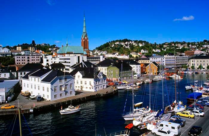 A vibrant marina scene is depicted, featuring a variety of boats docked along the waterfront. Colorful buildings line the shore, including a church with a distinctive clock tower in the background. The clear blue sky complements the lively atmosphere of the harbor.