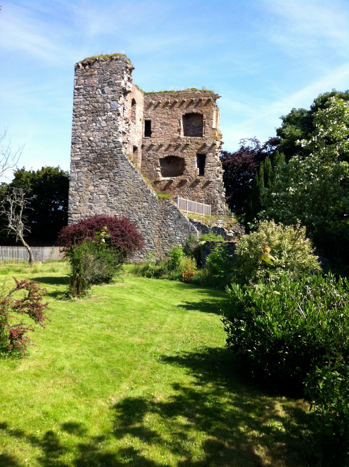 A historic stone tower, partially in ruins, rises amidst lush greenery. Surrounding gardens feature various shrubs and trees, creating a serene outdoor space. The remains of the structure showcase several open windows, revealing glimpses of the past against a clear blue sky.