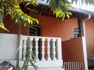 The entrance features a small balcony with a classic white balustrade, framed by lush greenery. The walls are painted in warm orange tones, creating a welcoming transition into the home. A doorway is visible, inviting guests into the interior space.