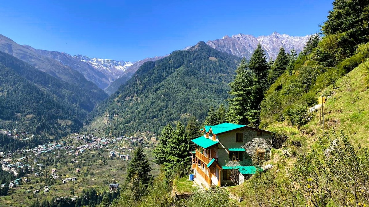 A traditional wooden and mud house is nestled in the mountains, showcasing a green roof. Surrounding lush greenery and tall trees lead to panoramic views of the deep valley below, with snow-capped mountains visible in the distance under a clear blue sky.