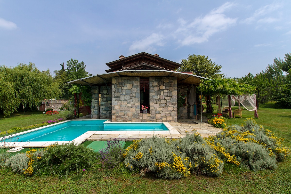 The villa's exterior showcases a stone façade surrounded by lush greenery. A private swimming pool is set into a paved area, complemented by vibrant flower beds. A shaded patio area offers seating options, enhancing the connection with the natural landscape.