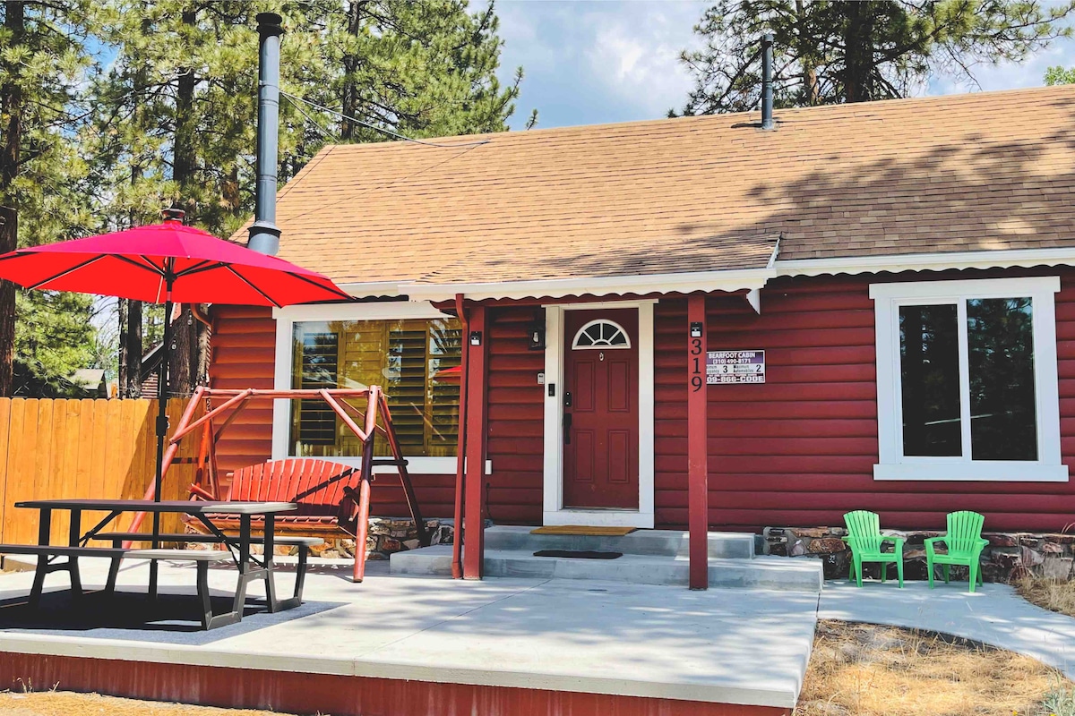 The outdoor space features a red picnic table under a vibrant umbrella alongside a cozy swing and green children's chairs. The cabin's exterior is a warm red, complemented by a stone pathway and wooden fencing, surrounded by tall trees.