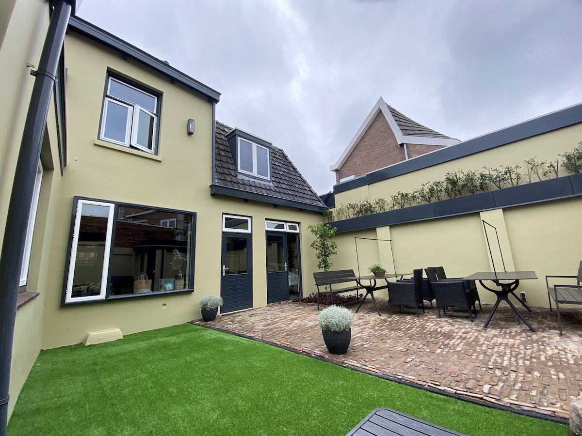 An outdoor seating area is visible, featuring a stone patio with several black dining tables and chairs. Lush green artificial grass borders the space, complemented by potted plants. The exterior of the building exhibits a modern design with large windows affording natural light.