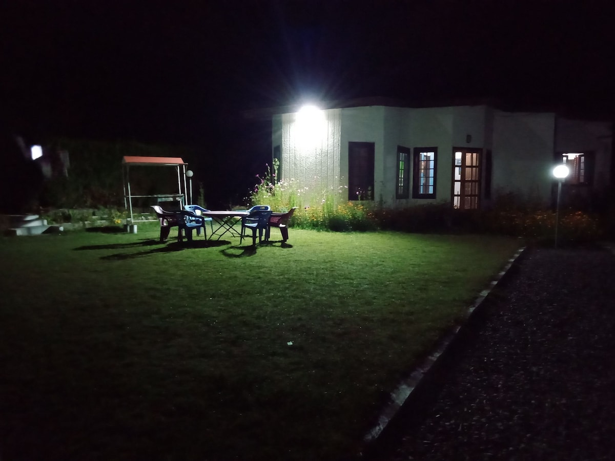 The night scene captures a well-lit garden surrounding a modest home. A table and two chairs are positioned on the grassy area, illuminated by a bright light source. The nearby flowers create a gentle backdrop against the building, which features multiple windows.
