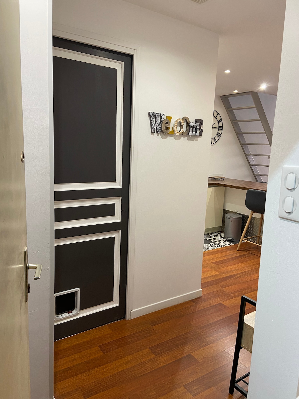 The entryway showcases a modern door with a unique pet access feature, accompanied by a welcoming sign that spells 'Welcome.' Light-colored walls contrast with the rich wooden floor, leading to an open living area visible in the background. A staircase ascends to the mezzanine.
