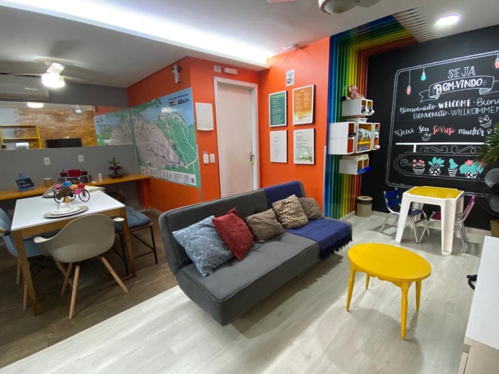 A modern shared space is highlighted with vibrant orange and green accents. A segmented seating area features a gray sofa with colorful cushions. A dining table is visible nearby, and decorative maps and posters adorn the walls, contributing to a welcoming atmosphere.