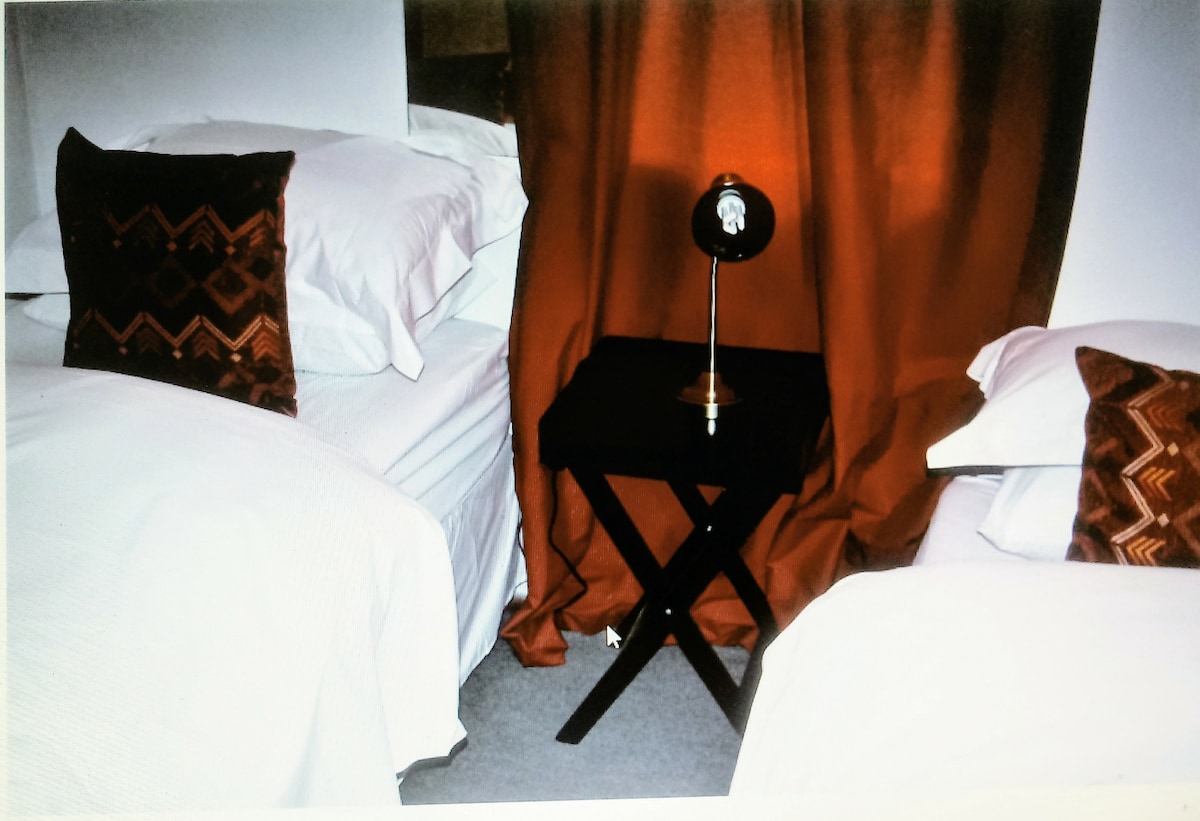 A cozy sleeping area features two neatly arranged beds, each topped with white bedding and decorative pillows. Between the beds, a small black side table supports an elegant lamp, while warm, orange curtains provide a touch of color to the serene space.