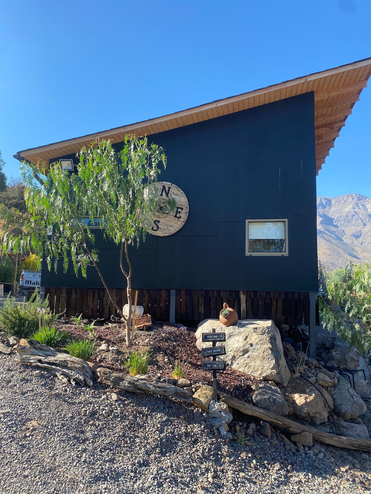 The exterior of a modern architectural structure is presented, featuring a dark façade accented by a large circular emblem. Nearby, a small tree and decorative stones are visible, with mountains rising in the background under a clear blue sky.