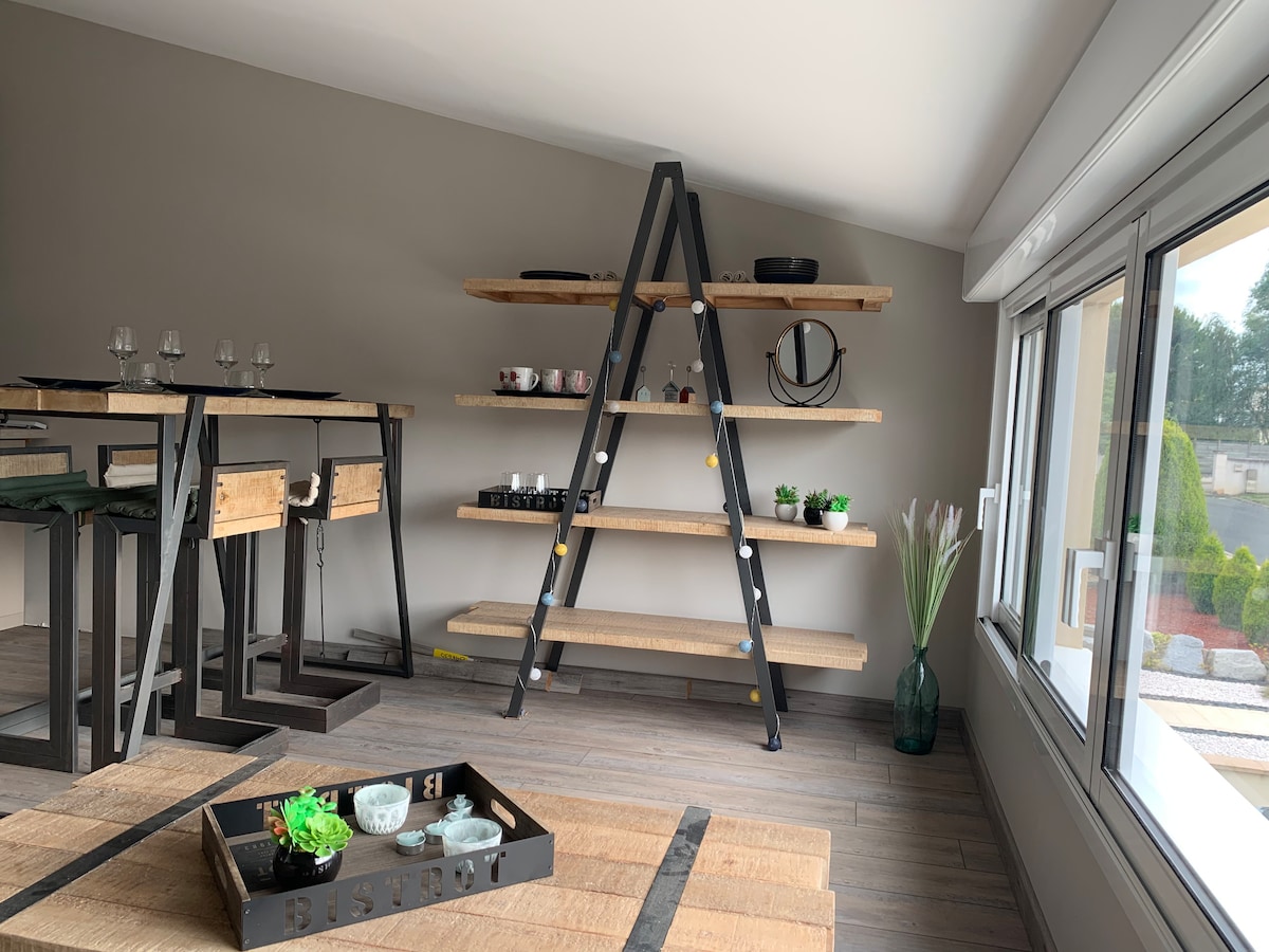 An open living area features a minimalistic shelving unit in a modern black and wood design, displaying decorative items. A dining table with four chairs is positioned nearby, and large windows allow natural light to illuminate the space.