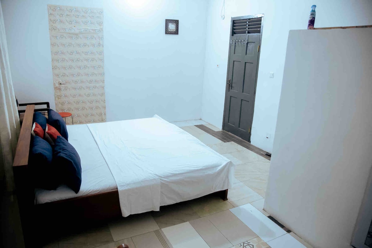A private room features a spacious bed with pillows against a neutral wall. A light-colored dresser stands nearby, and a simple door with a dark finish is visible. Light from a window brightens the space, contributing to a clean and uncluttered appearance.