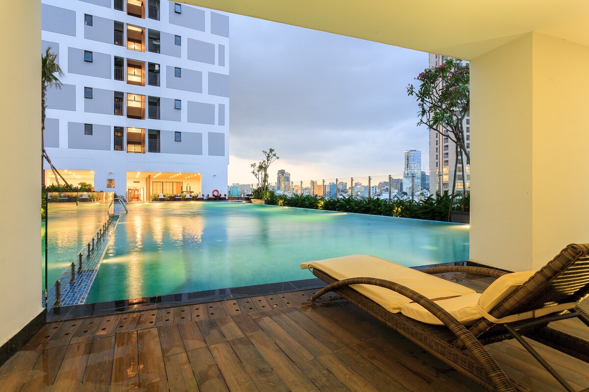 A modern swimming pool is showcased, featuring clear turquoise water and a spacious deck area with a lounge chair. Surrounding greenery and the city's skyline create a serene atmosphere. The scene is framed by a large opening that invites natural light into the space.
