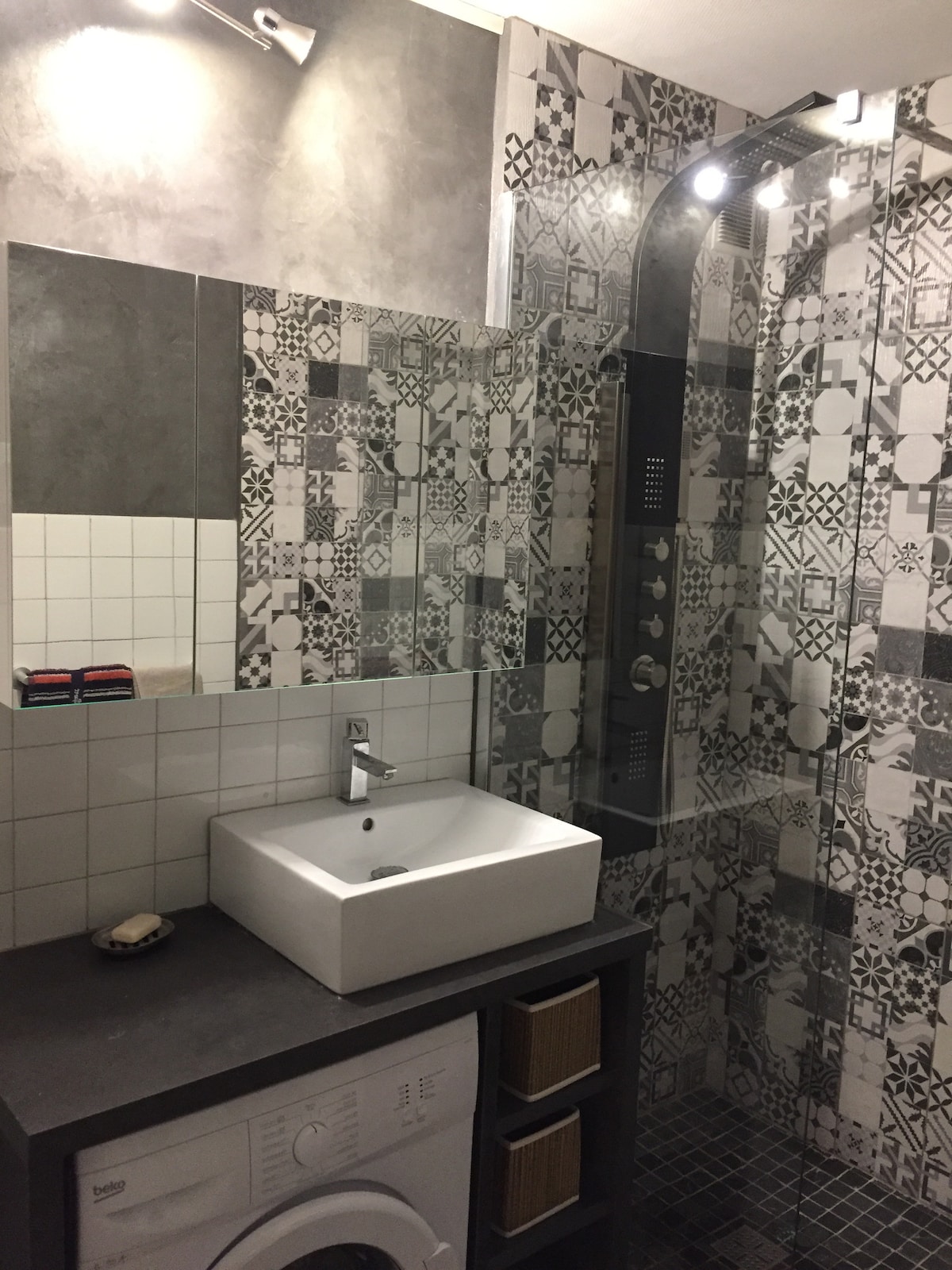 The renovated bathroom features modern tile work in black and white patterns. A glass shower with an overhead rain showerhead is visible, along with a sleek sink and ample counter space. A washing machine is located beneath the sink, providing convenience.