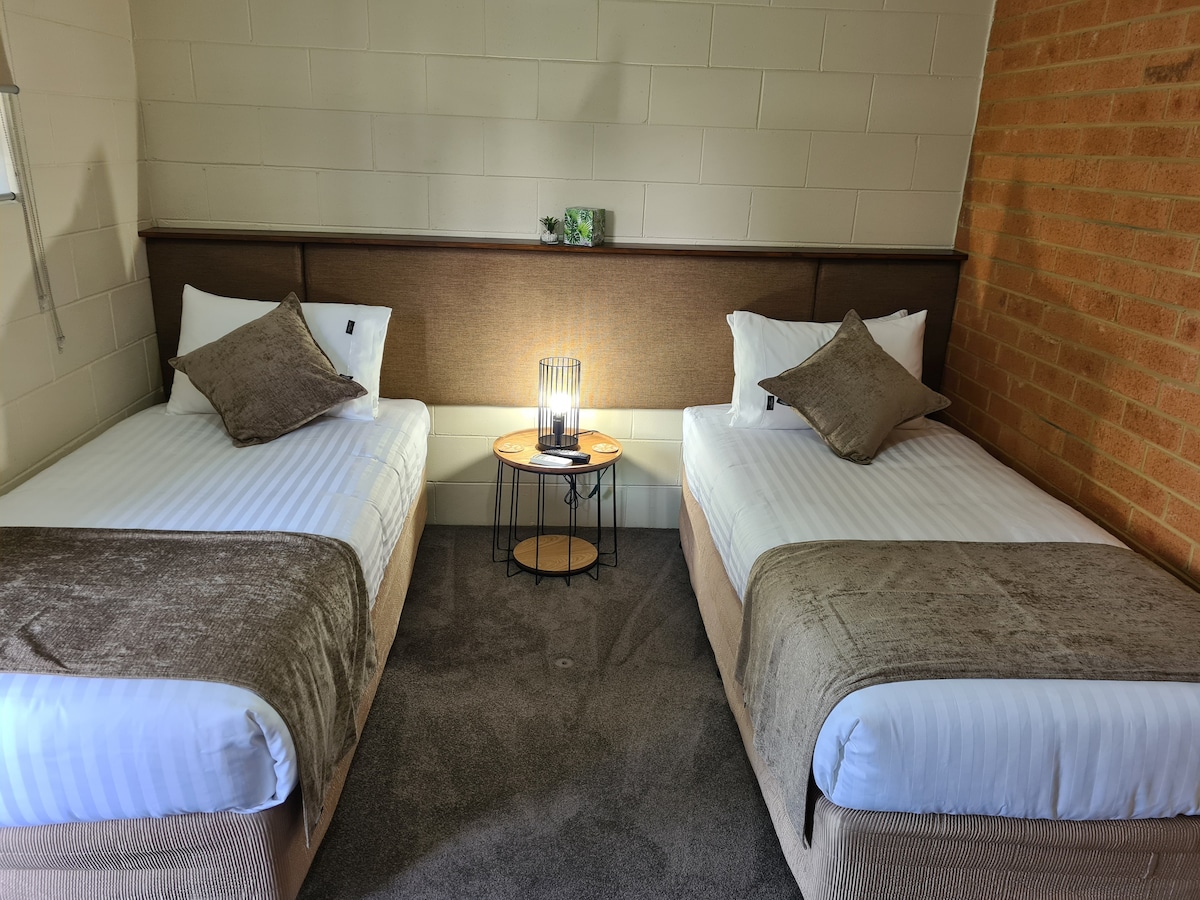 The sleeping area features two neatly made twin beds, each adorned with soft blankets and decorative pillows. A small round table is situated between the beds, accompanied by a softly glowing lamp. The walls are a mix of light-colored paint and exposed brick.