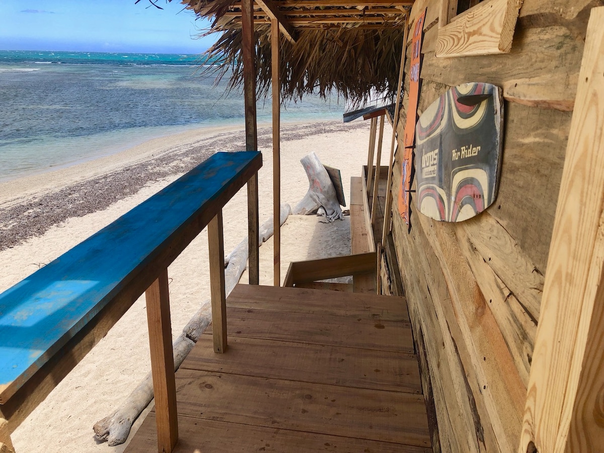 A wooden deck sits adjacent to the white sandy beach, featuring a long blue bench under a thatched roof. To the side, surfboards are displayed on the rustic wooden wall, and the calm turquoise waters can be seen in the background.