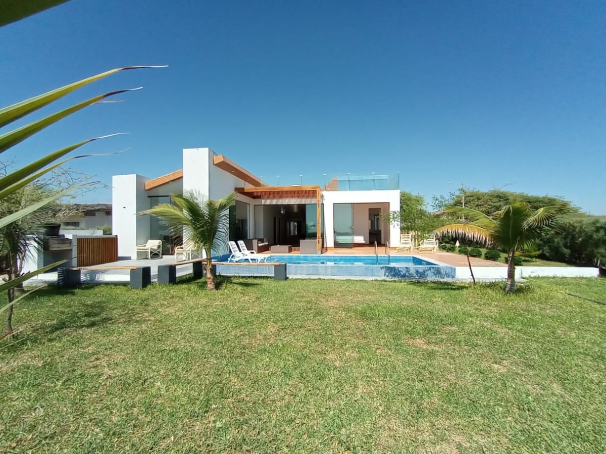 A modern eco-sustainable house is surrounded by green grass, featuring an inviting private pool. Large windows offer views of the living area and terrace. Palm trees provide shade, enhancing the serene outdoor space, ideal for relaxation.