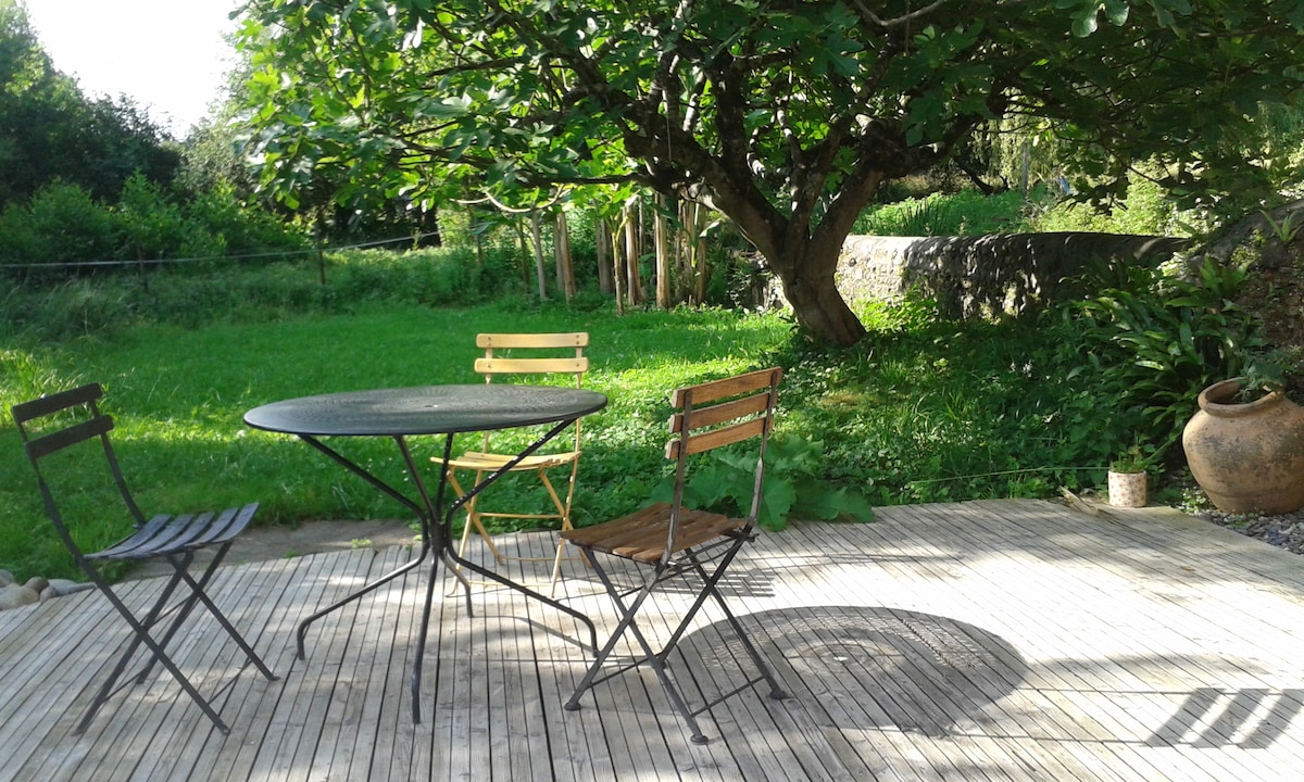 An outdoor seating area features a round black table surrounded by two metal chairs and one wooden chair. A lush green garden is visible in the background, with a mature tree providing shade. The wooden deck offers a natural setting for relaxation.