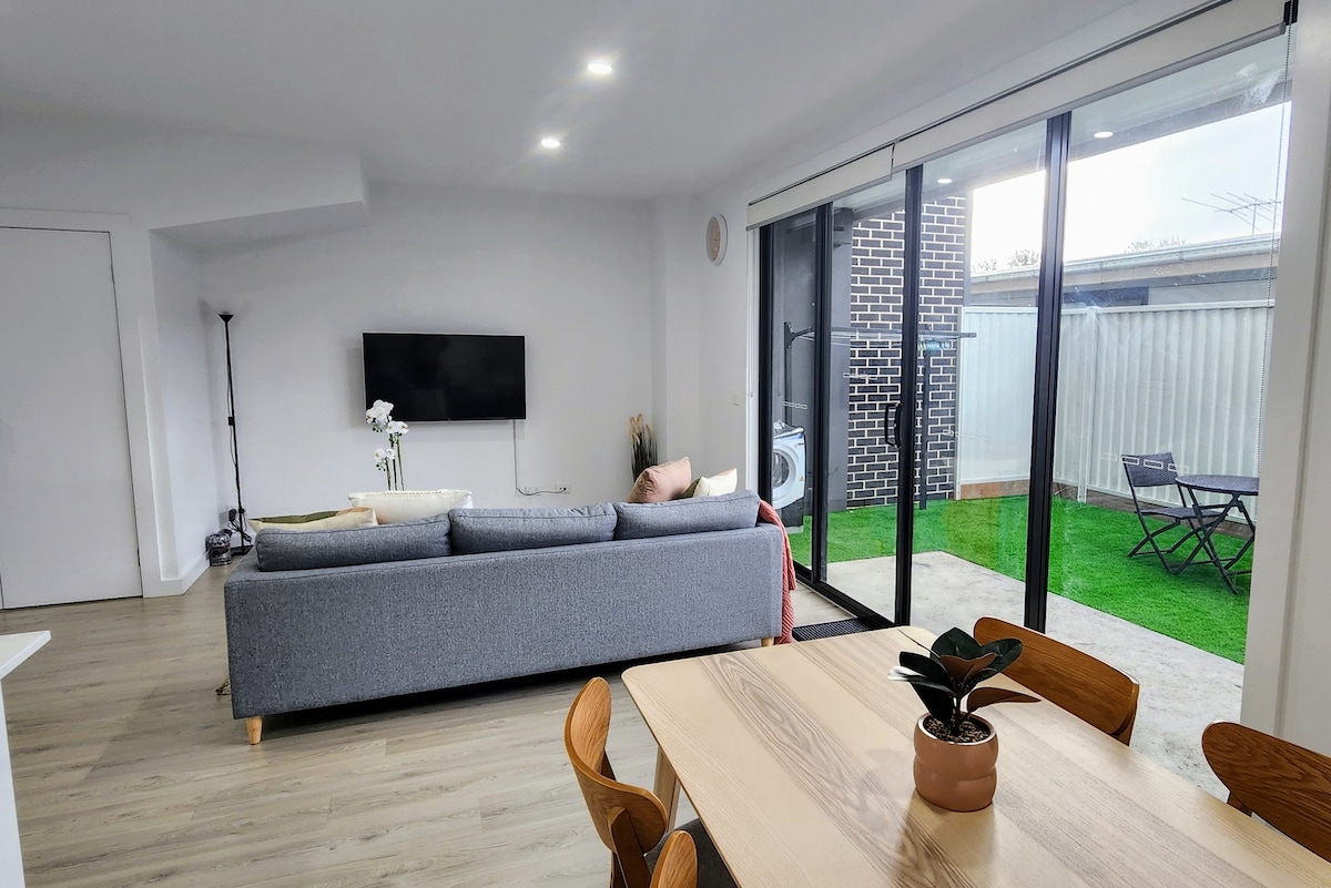 A modern living area showcases a comfortable gray sofa facing a flat-screen TV mounted on the wall. Large glass doors provide access to a private courtyard with a small outdoor dining set, while a dining table with wooden chairs occupies the foreground.