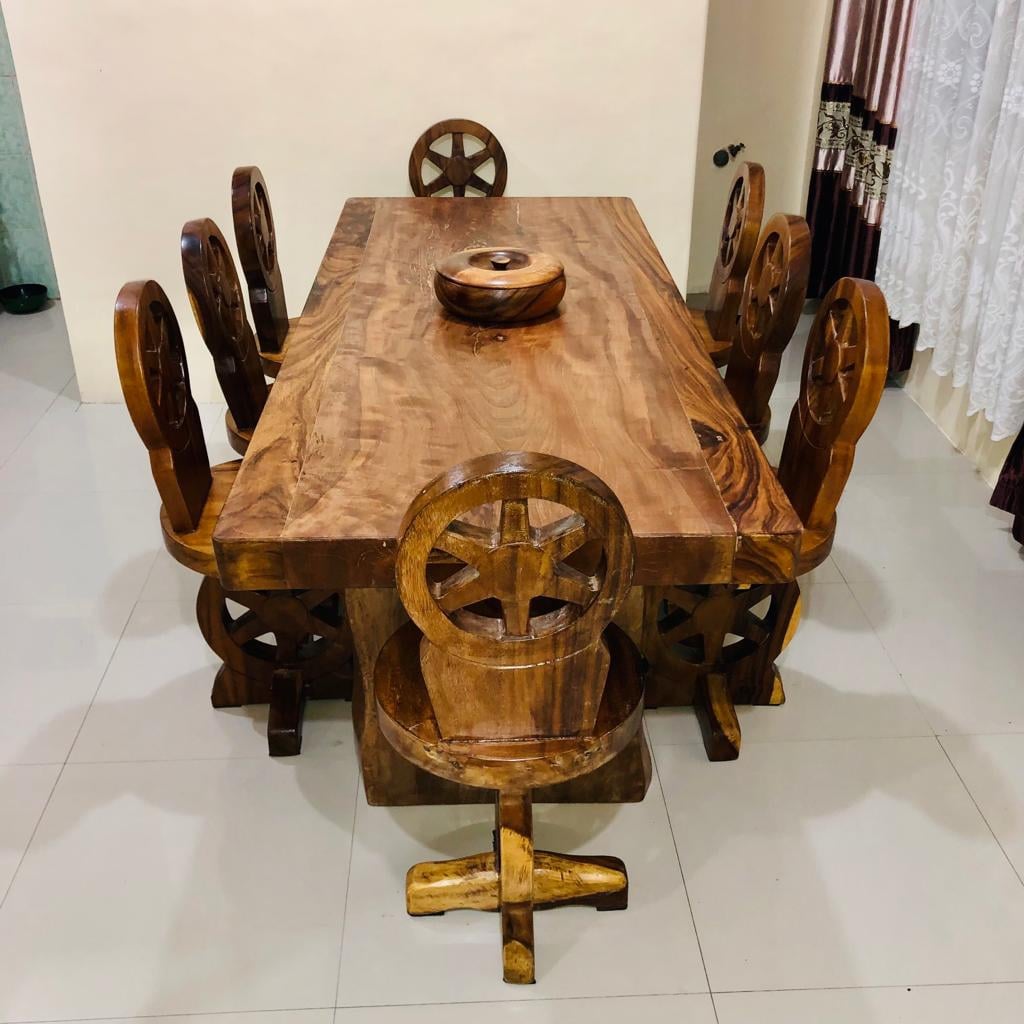 A large wooden dining table features a smooth surface and is accompanied by eight intricately designed chairs. Each chair displays a wheel motif, creating a cohesive aesthetic. Natural grain patterns are visible in the wood finish, and the table's center holds a circular wooden accessory.