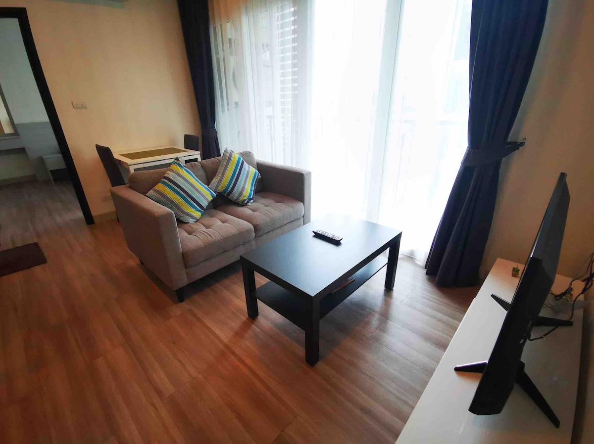 A cozy living area features a comfortable sofa adorned with colorful striped cushions, complemented by a sleek black coffee table. Natural light streams through large glass doors, creating a bright atmosphere. A television is positioned on a white media console against the wall.