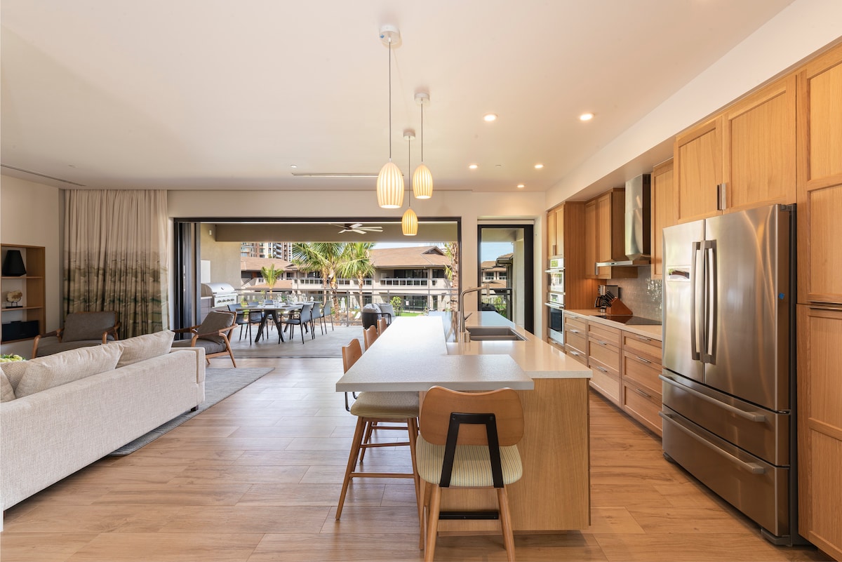 A modern kitchen area is presented with wooden cabinetry and stainless steel appliances. A spacious island with seating is visible, along with large sliding doors that open to the outdoor dining area, seamlessly integrating indoor and outdoor spaces.