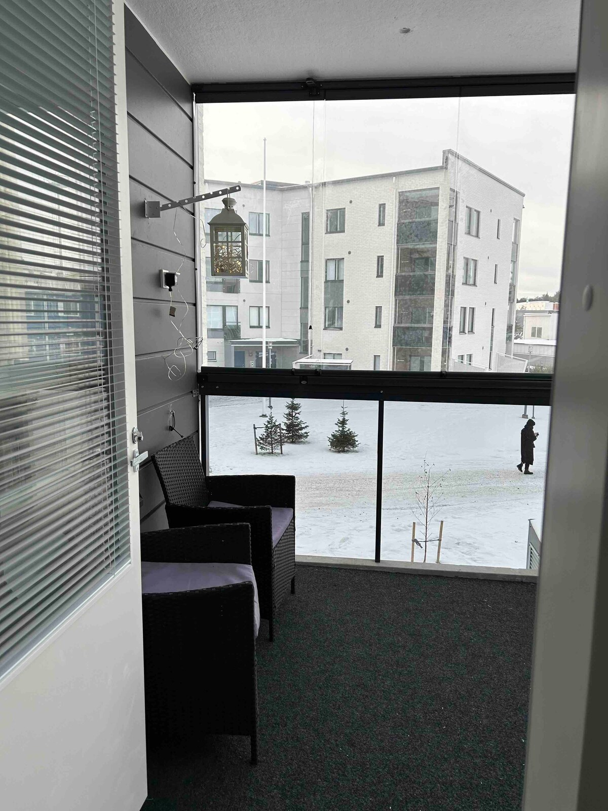 A covered balcony area is displayed with two black wicker chairs, offering a cozy space to enjoy the outdoors. A green carpet complements the setting, while large glass panels provide an unobstructed view of the snowy landscape and surrounding buildings.