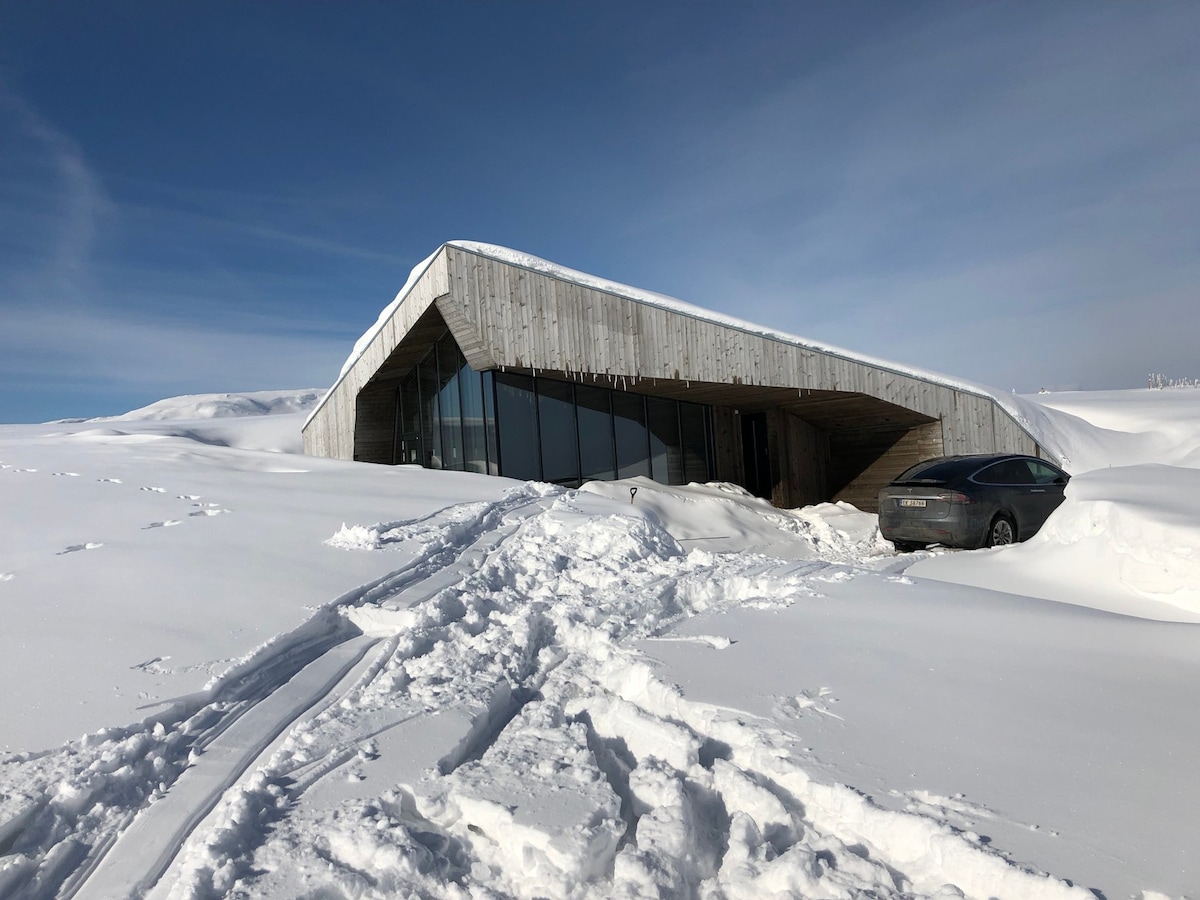 The modern cabin, designed with a sloping roof, is partially covered in snow, showcasing its unique architecture. Large windows reflect the clear blue sky and surrounding snow-covered landscape. A parked car is visible near the entrance, indicating convenient access.