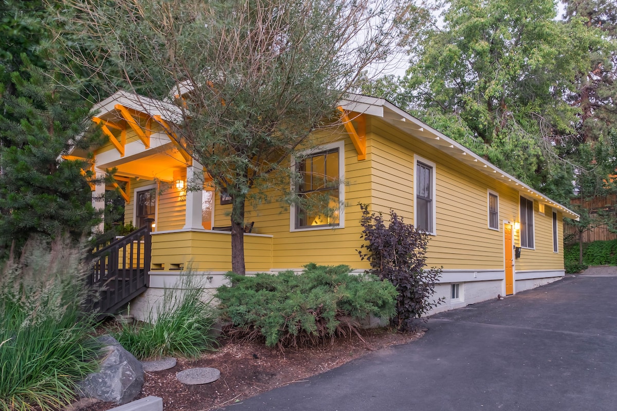 The exterior of a historic three-bedroom home is showcased, featuring a warm yellow façade with white trim. A welcoming entrance is framed by trees and shrubs. An asphalt driveway provides access to the property, which is nestled in a lush, green environment.