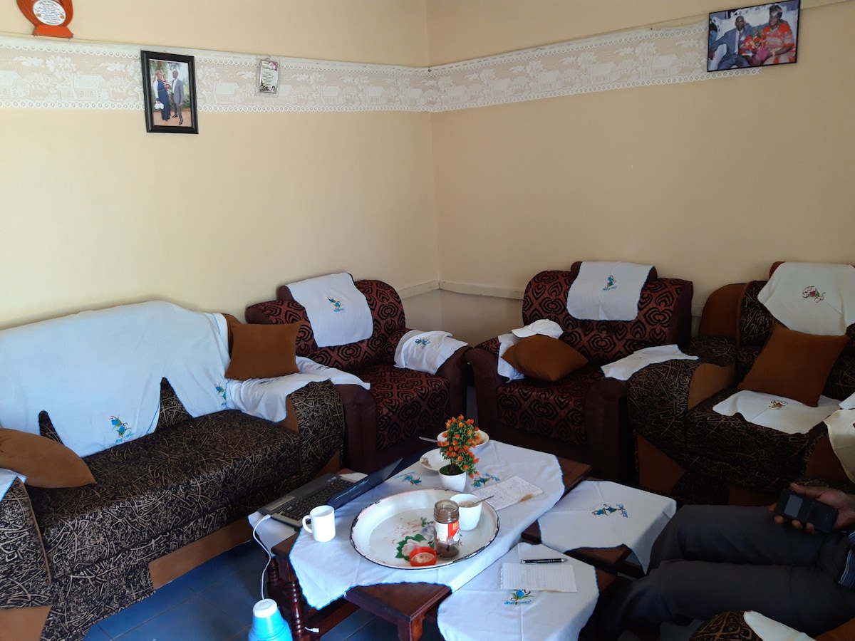 A furnished living room features two patterned sofas with decorative cushions. A round table is set with a tray, candle, and small plant. Wall decor includes framed photographs, creating a comfortable space for relaxation.