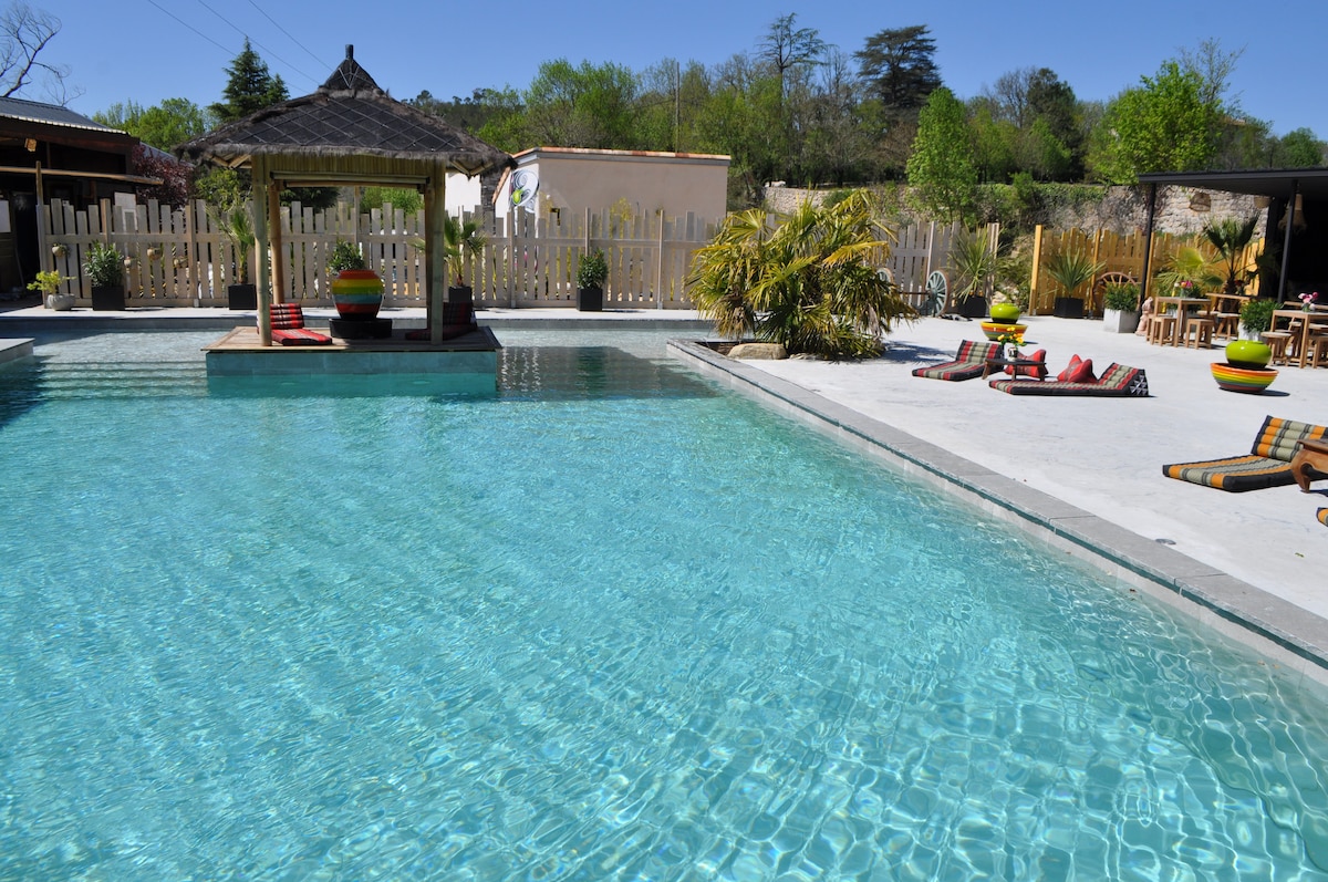 A spacious outdoor swimming pool reflects clear blue skies, surrounded by palm trees and lounge areas. A shaded gazebo is positioned at the pool's center, while colorful lounge chairs are placed along the water's edge, offering a peaceful space for relaxation.