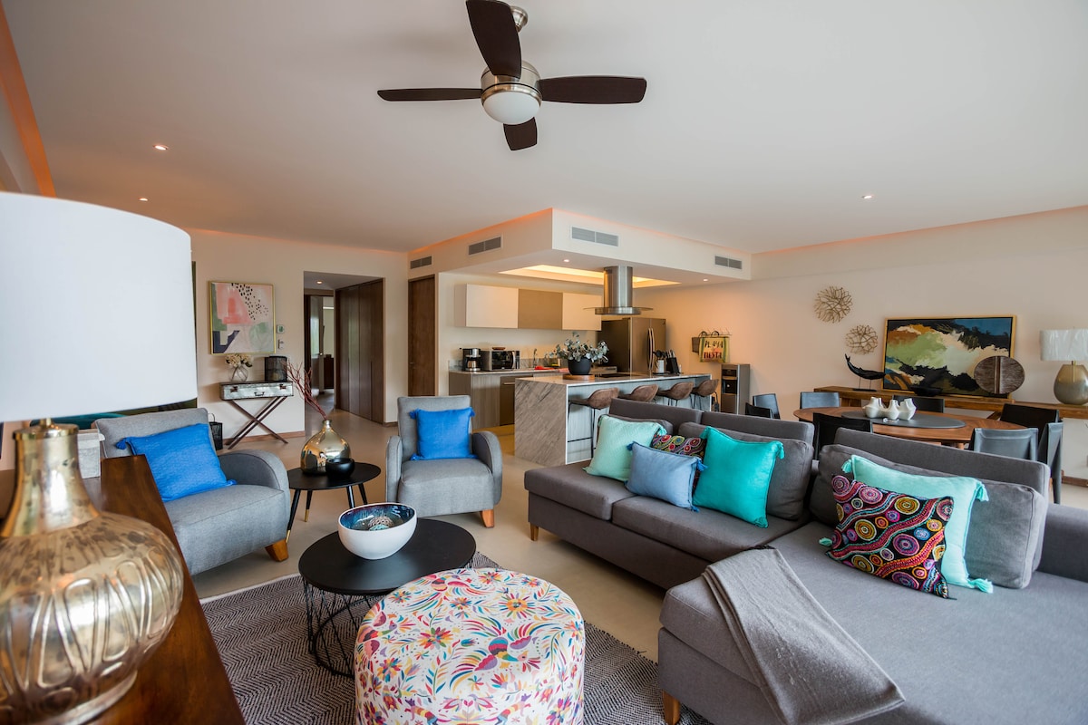 A modern living area features comfortable seating arranged around a central coffee table. Accent cushions in vibrant colors complement the neutral upholstery. A ceiling fan above contributes to airflow, while an open kitchen area is visible in the background, enhancing the spacious ambiance.