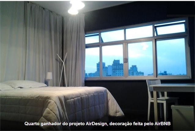 A comfortable sleeping area features a spacious bed with a light quilt, positioned next to large windows that provide a view of the city skyline. A small table and chairs are visible, creating an inviting space for meals or work.