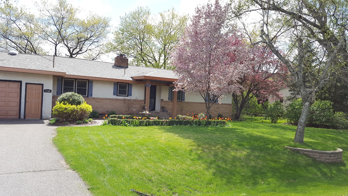 A well-maintained bungalow is set in a lush green landscape, featuring colorful flower beds and two blooming trees. A wide driveway leads to the garage, while manicured grass surrounds the entryway, inviting visitors to the front porch.