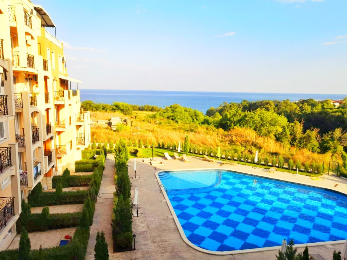 A large outdoor swimming pool is framed by neatly arranged garden beds and sun loungers. The pool's vibrant blue tiles reflect the clear sky. In the background, trees and greenery extend towards the coastline, showcasing a panoramic view of the sea.