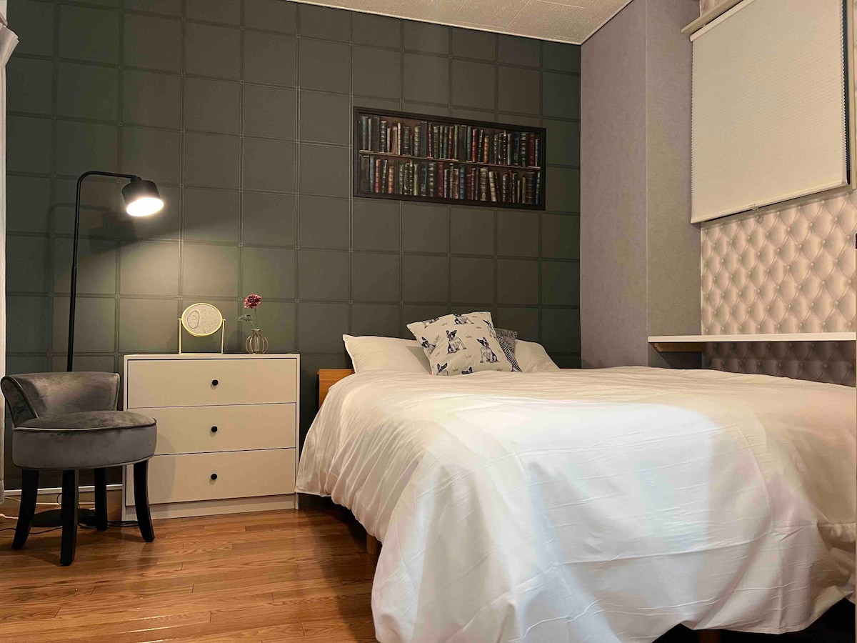 A bedroom is presented with a spacious bed covered in white linens. A stylish lamp illuminates the room, while a small flower vase adds a touch of decor. A dresser with drawers and a reading chair are positioned nearby, with patterned wall art above the bed.