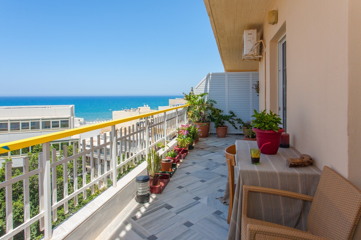 A furnished balcony with a table and chairs overlooks the sea, surrounded by potted plants. The tiled floor is complemented by a railing providing safety and a view of the blue ocean and nearby buildings.