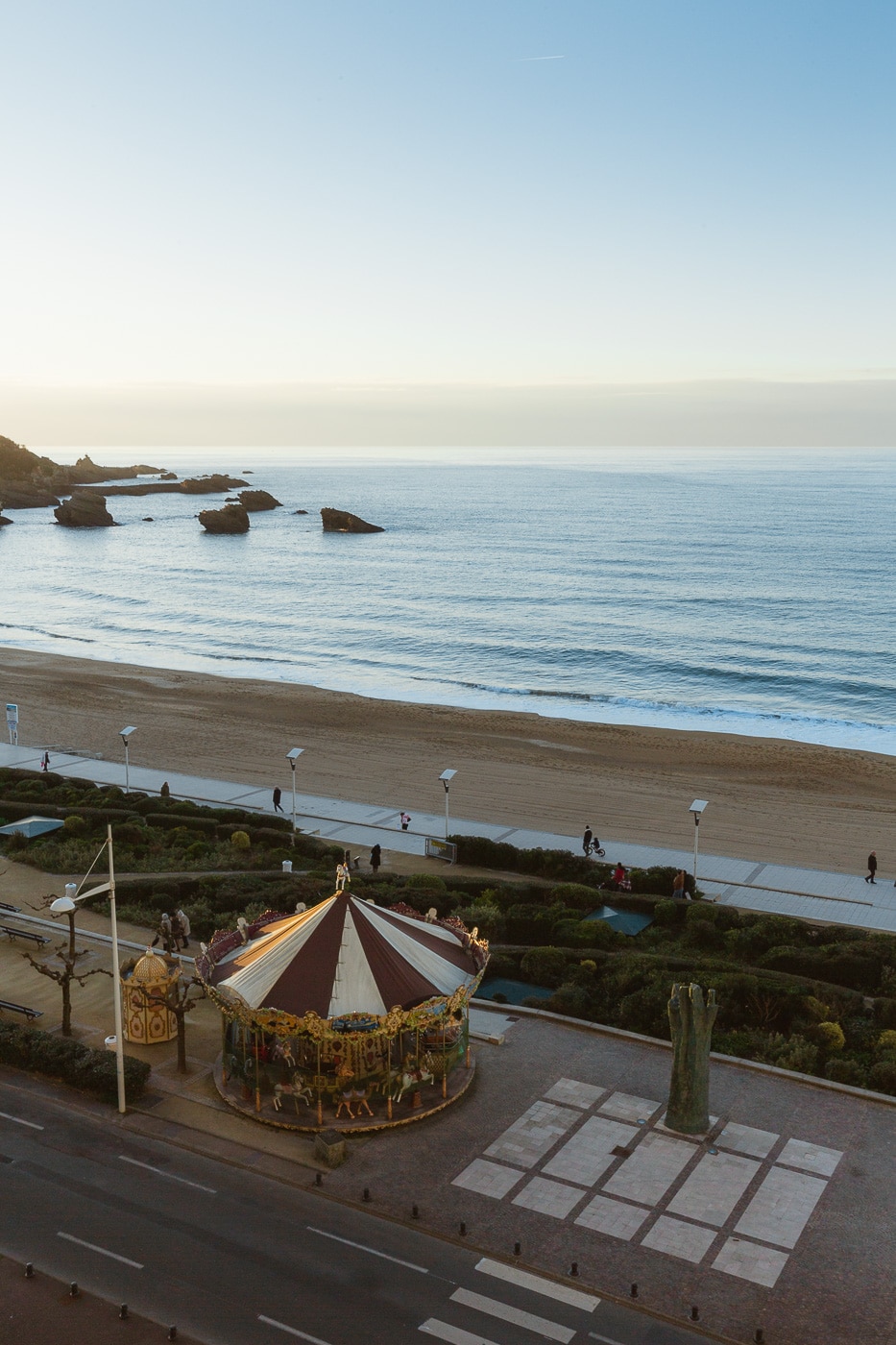 An ocean view shows a sandy beach bordered by gentle waves, with rocky formations in the distance. A colorful carousel occupies the foreground, surrounded by landscaped greenery and pathways. The sky transitions from soft blue to warm hues as the day progresses.