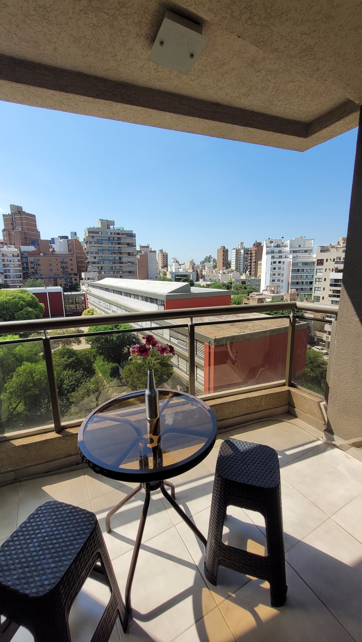 A balcony is featured, offering a view of the city with buildings in the background and greenery below. A round table is set with a vase containing flowers, accompanied by two high-backed chairs. Natural light illuminates the space.