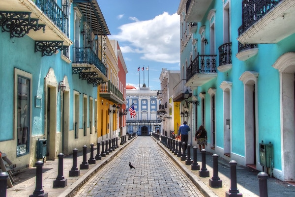 A vibrant street view showcases colorful buildings in shades of blue, yellow, and white. A cobblestone pathway leads towards a central plaza with flags prominently displayed. Decorative street lamps line the sidewalk, while pedestrians stroll leisurely, creating a lively atmosphere.
