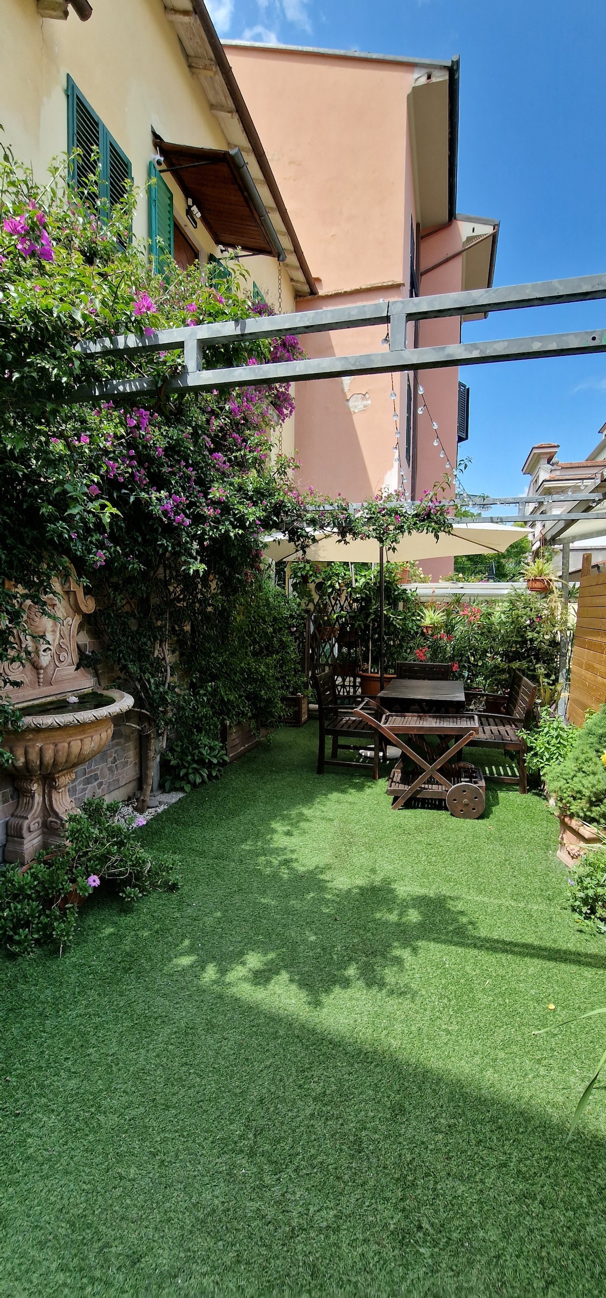 A private garden area is available, featuring lush green grass and vibrant flowers climbing trellises. A wooden dining table, surrounded by chairs, invites outdoor meals. A calming water feature is positioned along the wall, contributing to the serene atmosphere.