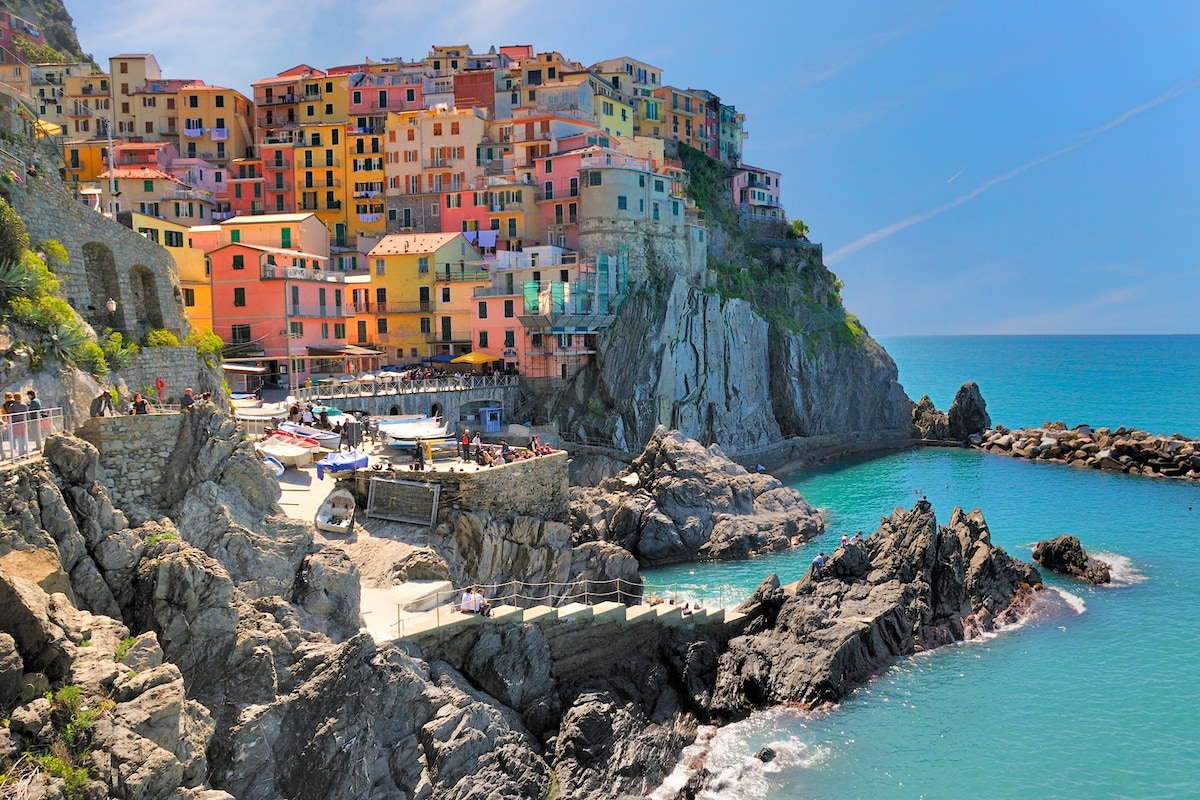 A coastal village is nestled against rocky cliffs, featuring colorful buildings with varied facades. The clear blue water reveals boats anchored along the shore, creating a tranquil scene. Natural rock formations and gentle waves enhance the picturesque setting, inviting exploration of the surrounding landscapes.